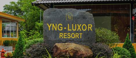 Un letrero de piedra con el nombre "YNG-LUXOR RESORT" en letras amarillas, rodeado de follaje y un edificio con tejado de tejas.