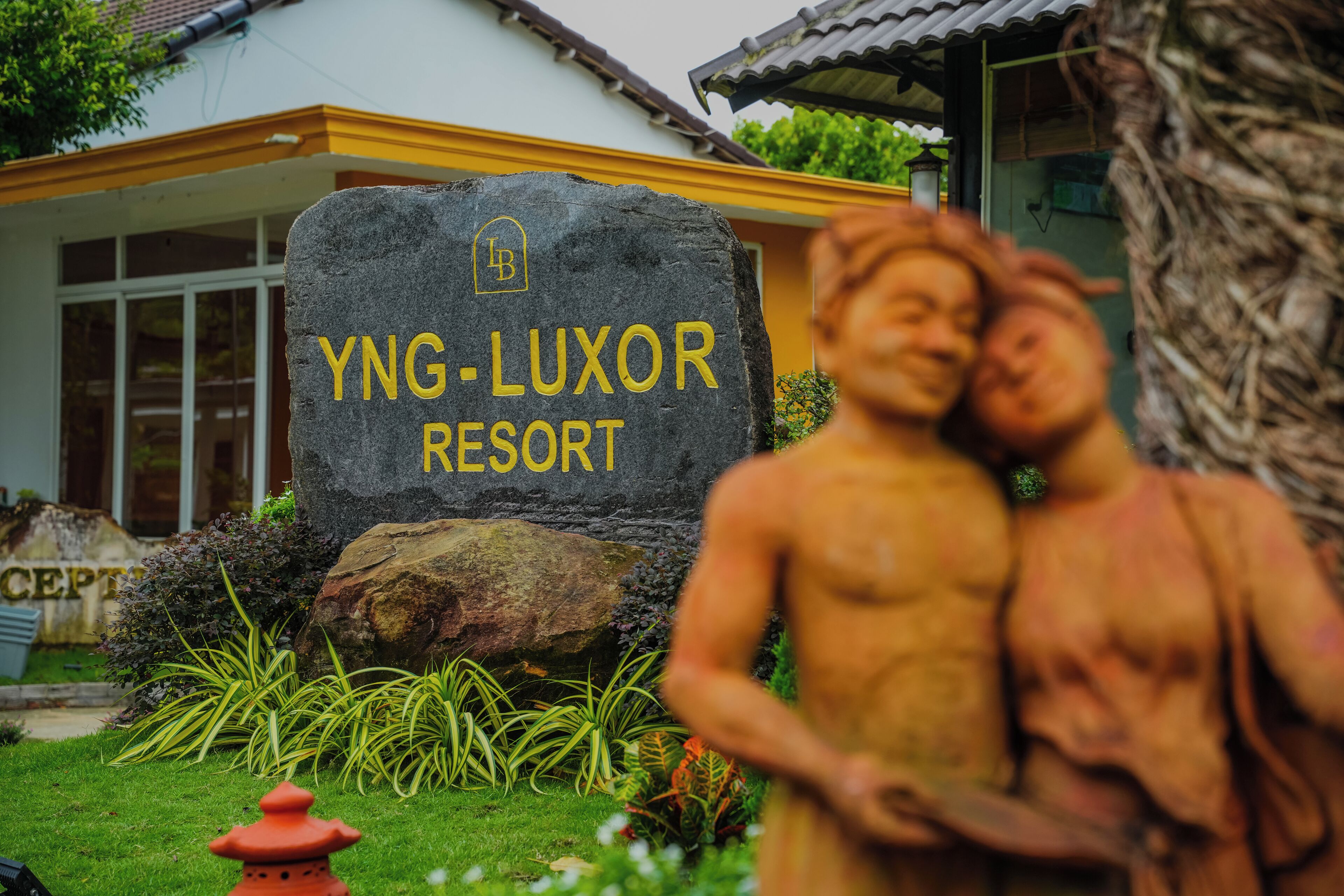 Un cartello in pietra con la scritta "YNG-LUXOR RESORT", circondato da vegetazione e una statua.