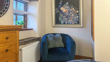 Une pièce avec un fauteuil bleu, une commode en bois, un radiateur et un tableau représentant un paon, encadré, accroché au mur.