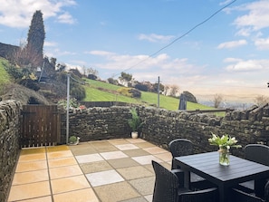 Une terrasse avec un sol en pierre, une table, des chaises, un pot de fleurs et un mur en pierre.
