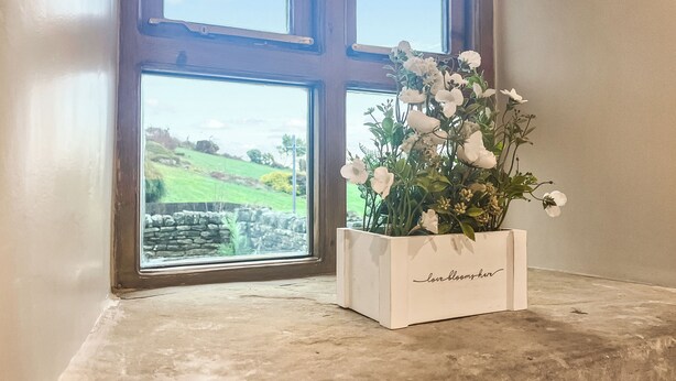 Une fenêtre donnant sur des champs verdoyants et des murs de pierre, une jardinière blanche sur laquelle est inscrit « L’amour fleurit ici », et un sol en béton.