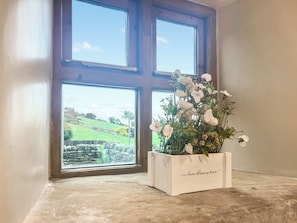 Une fenêtre donnant sur des champs verdoyants et des murs de pierre, une jardinière blanche sur laquelle est inscrit « L’amour fleurit ici », et un sol en béton.