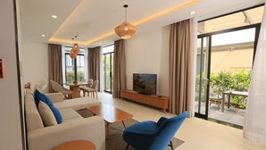A modern living room with a sofa, blue armchair, coffee table, TV, and sliding glass doors to an outdoor area. - OCEAN BREEZE VILLA CAM RANH (Cam Lam)
