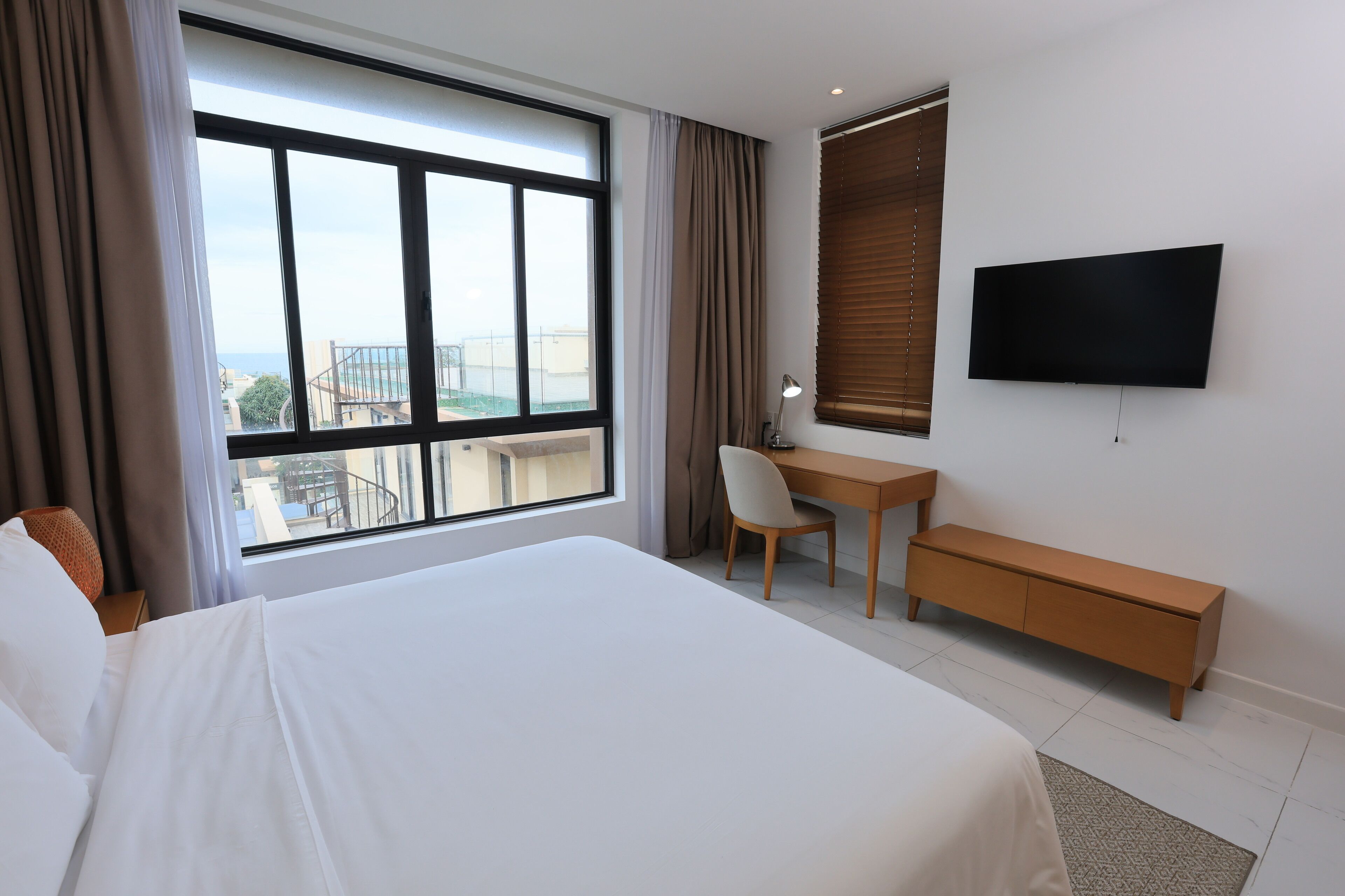 A hotel room with a large bed, a desk, a chair, a TV, and a view of the city through the window.