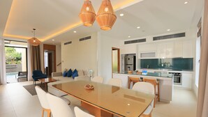 A modern living room with a glass-top dining table, white sofas, and a kitchen in the background. - OCEAN BREEZE VILLA CAM RANH (Cam Lam)