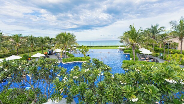A large swimming pool surrounded by lush greenery and palm trees, with a view of the ocean in the background. - OCEAN BREEZE VILLA CAM RANH (Cam Lam)