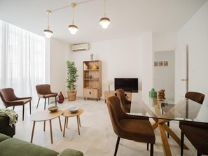 A modern living room with a glass dining table, brown cushioned chairs, a wooden bookshelf, a television, and a large window with sheer curtains.