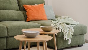 A green sofa with two bowls on a small round table, a green throw blanket, and two pillows in orange and blue.