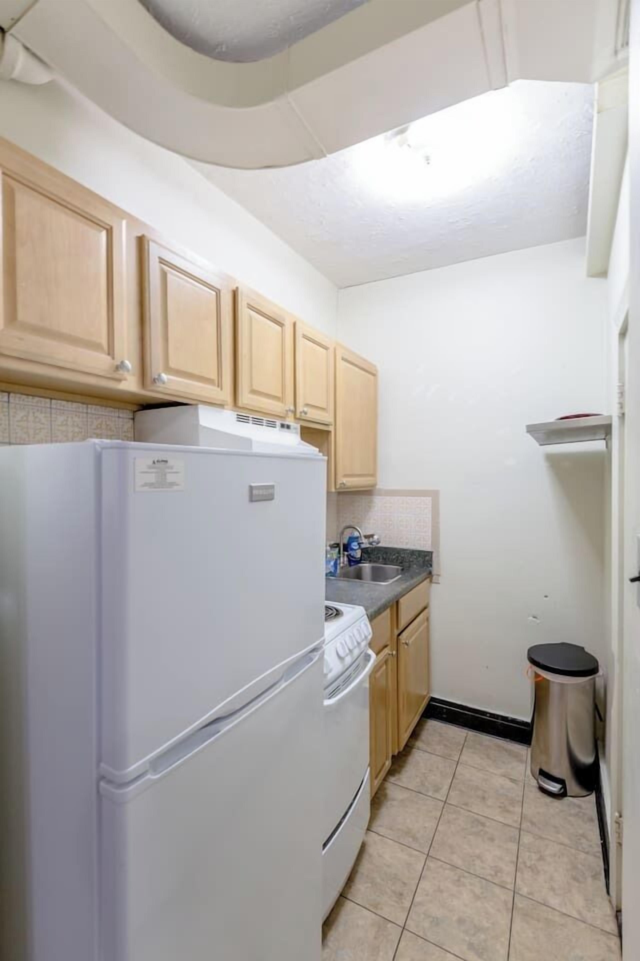 A compact kitchen with wooden cabinets, a white refrigerator, a stove, a sink, and a trash bin.