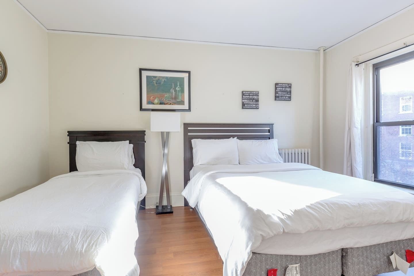 Ett hotellrum med två sängar, en golvlampa, en inramad tavla på väggen och ett fönster med utsikt över byggnader.