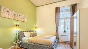 Une chambre à coucher avec une tête de lit capitonnée, une table de chevet ornée d’une plante, une grande fenêtre avec des rideaux et une œuvre d’art accrochée au mur.