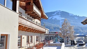 A building with a wooden balcony, a snow-covered landscape, and a mountain in the background. - Holiday Apartment with a spa next to the gondola (Brixen im Thale)
