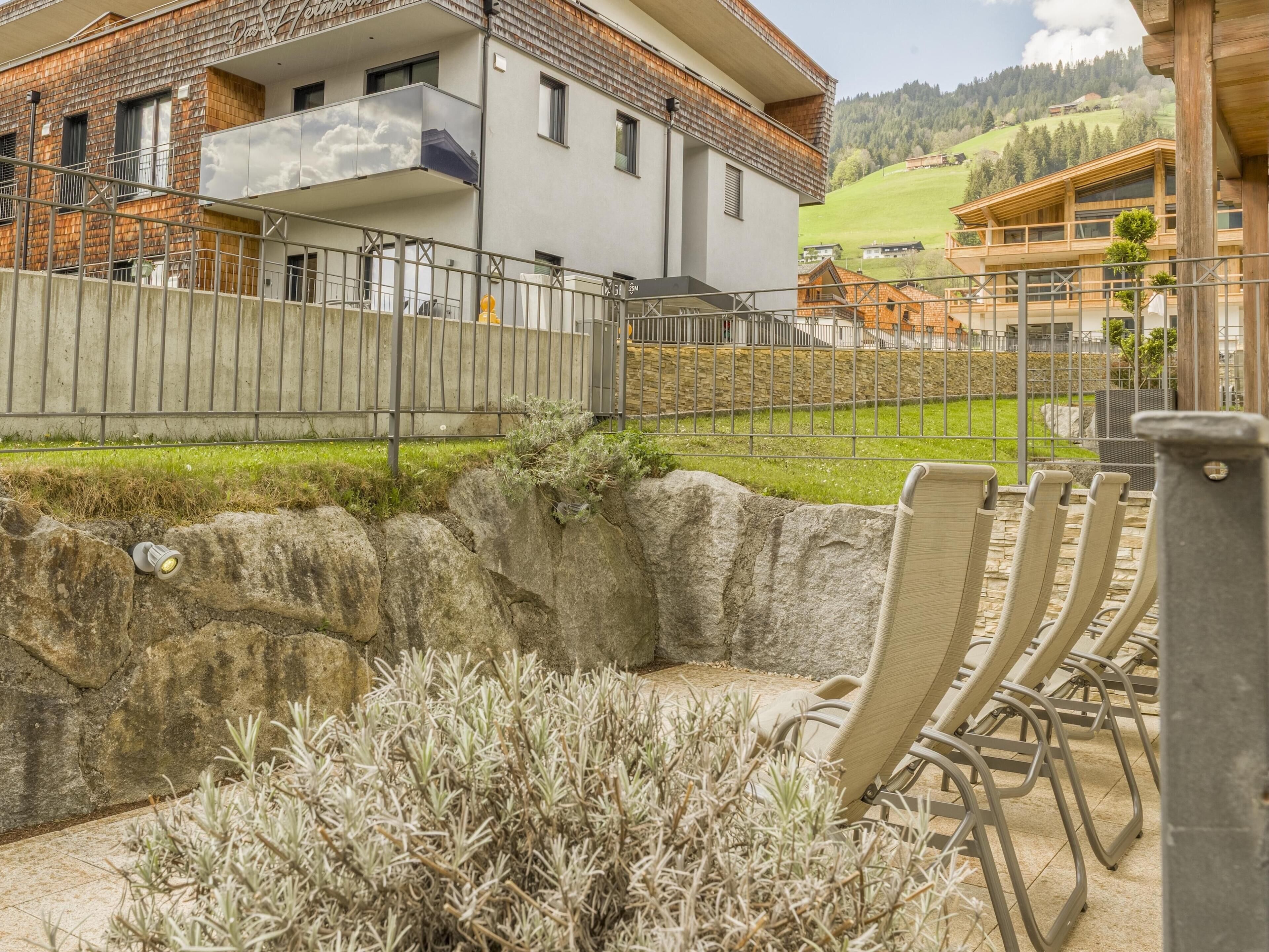 Un edificio moderno con un balcón, un muro de piedra de contención y un área ajardinada con sillas.