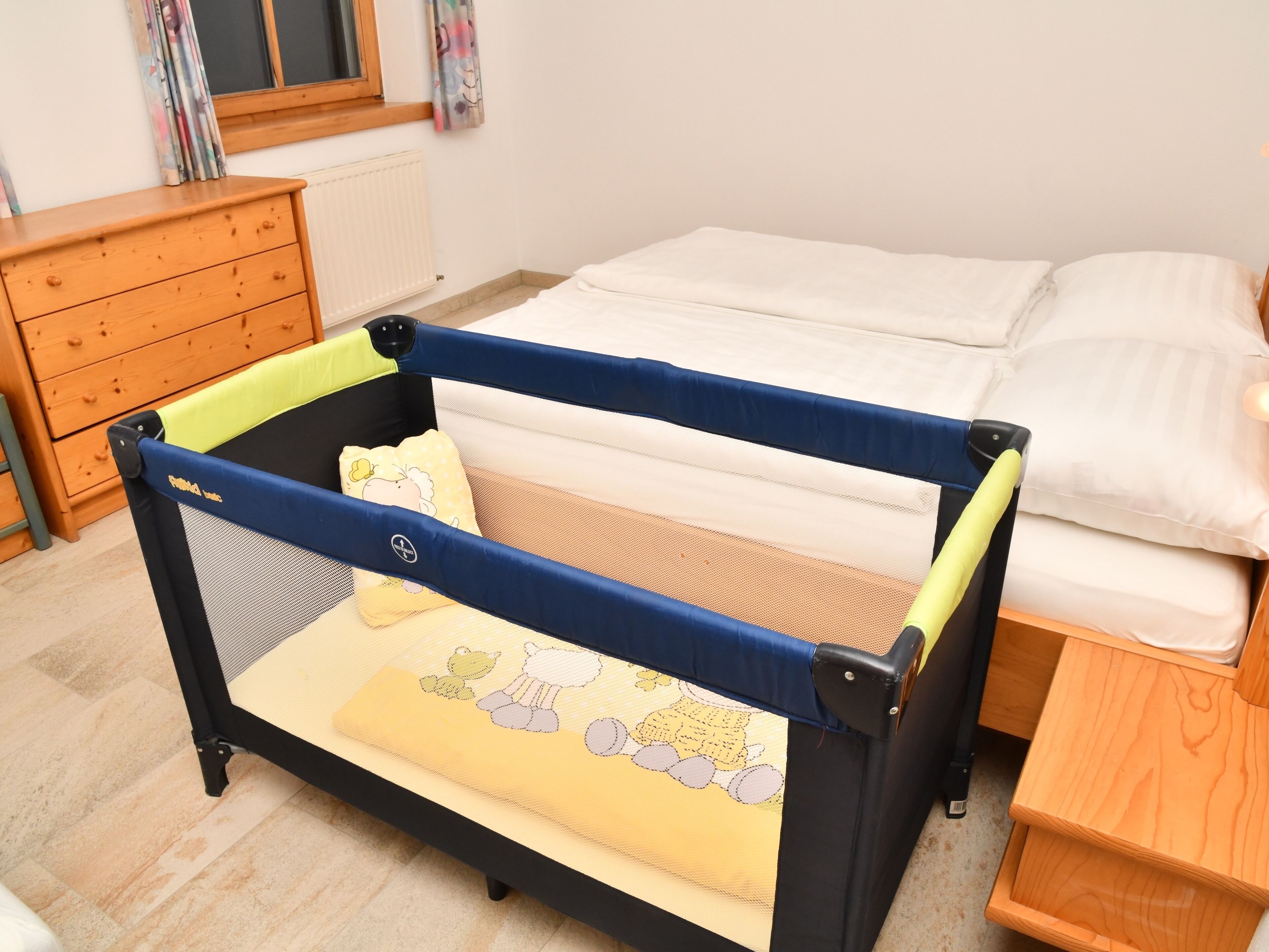 A child's playpen with a yellow cushion and cartoon character design, a wooden dresser, and a bed with white bedding.