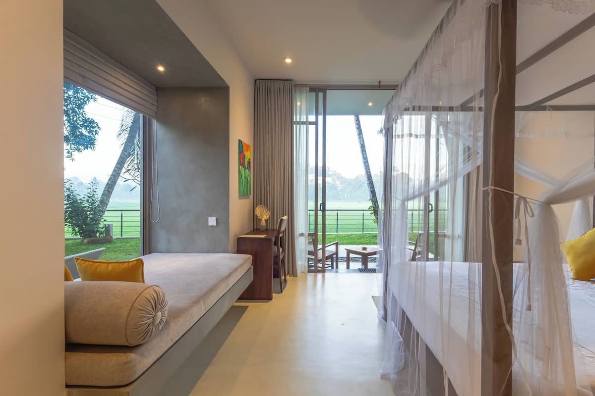 Quarto de hotel moderno com cama, escrivaninha, cadeira e vista para uma paisagem verde através de grandes janelas.