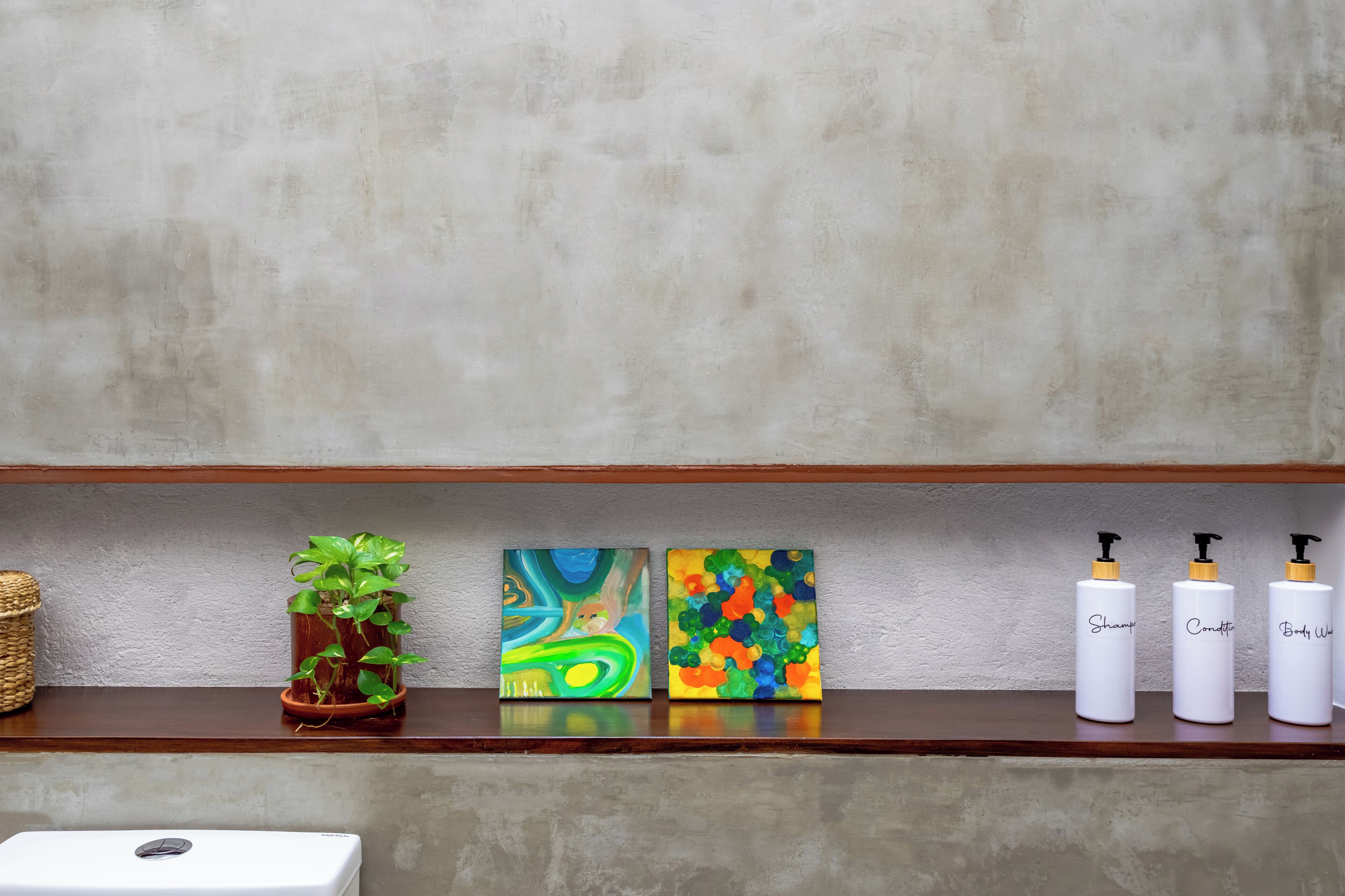 Uma prateleira com uma planta em vaso, duas pinturas abstratas e três garrafas brancas com bombas pretas.