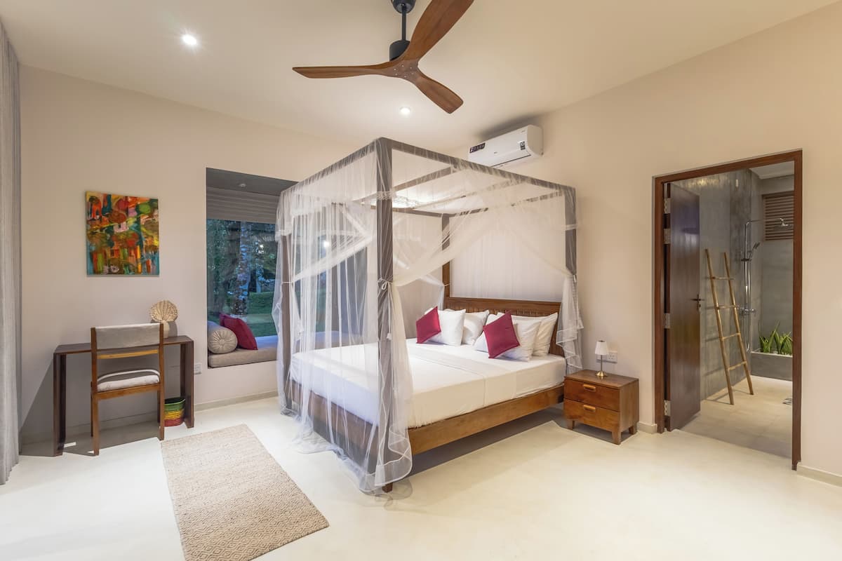 Quarto com cama de dossel, ventoinha no teto, ar condicionado, mesa de cabeceira, cadeira, janela com vista e porta para área do chuveiro.