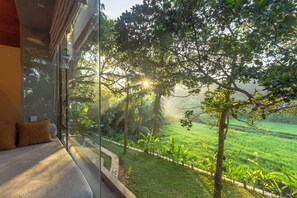 A room with a large window overlooking a lush green landscape, a bed with pillows, and a view of a tree-lined path. - Ona Garden Villa  (Galle)