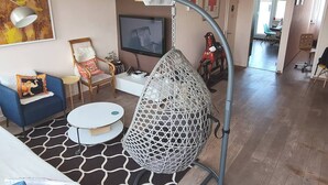Una sala de estar con un sofá azul, una mesa de centro blanca, una silla colgante, un televisor y una zona de comedor al fondo.