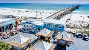 Un hotel frente a la playa con un muelle que se adentra en el océano, sillas de playa y un edificio con techo de paja.