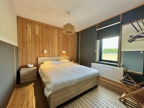 A bedroom with a wooden bed, bedside tables, a hanging light fixture, a window with a view, and a radiator.