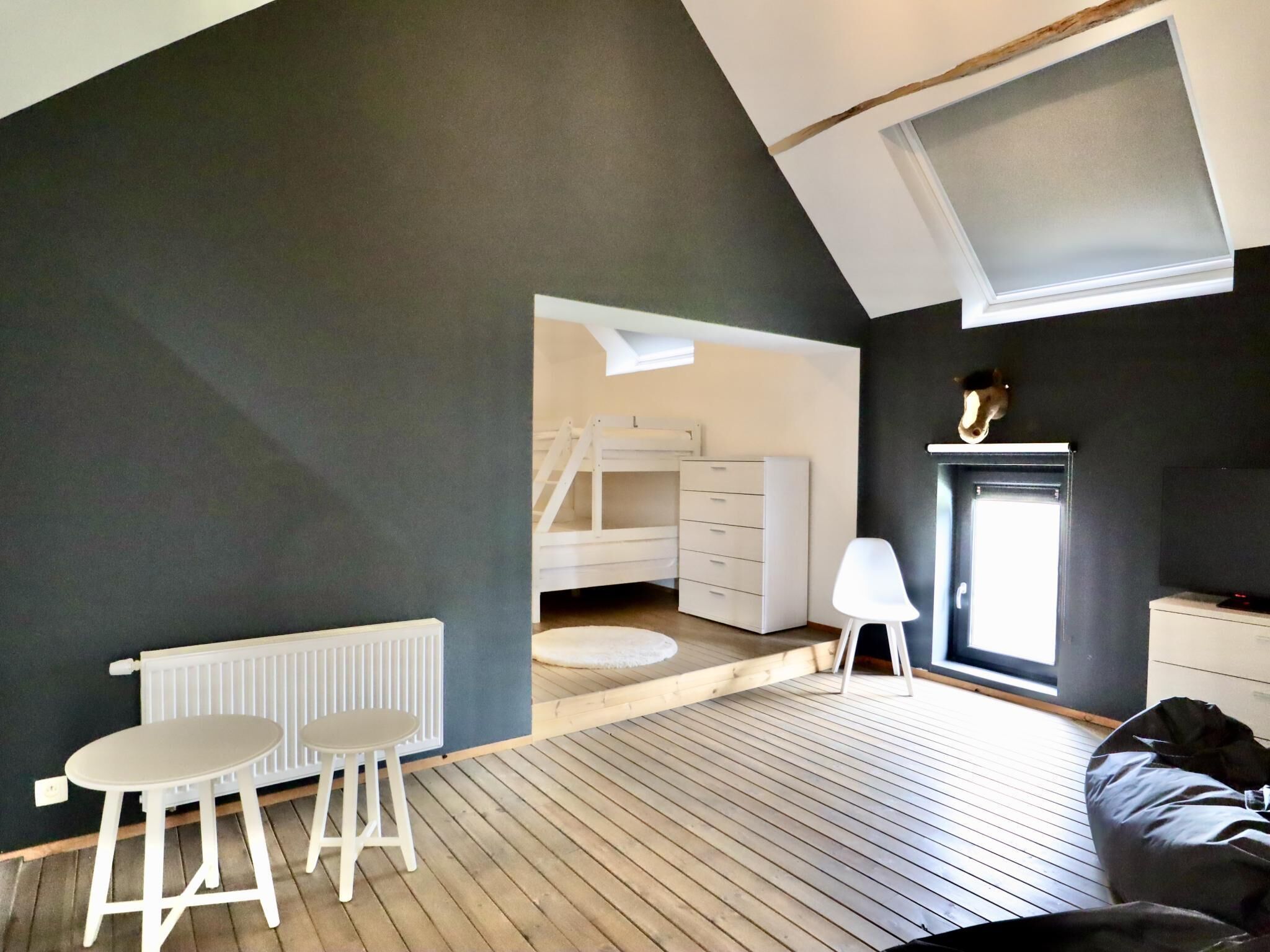 Une pièce avec un plafond en pente, un radiateur blanc, une petite table blanche, une chaise blanche, une commode blanche, un plancher en bois et une fenêtre.
