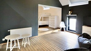 Ein Zimmer mit schrägverdeckter Decke, einem weißen Heizkörper, einem kleinen weißen Tisch, einem weißen Stuhl, einer weißen Kommode und einem Holzboden, sowie einem Fenster.