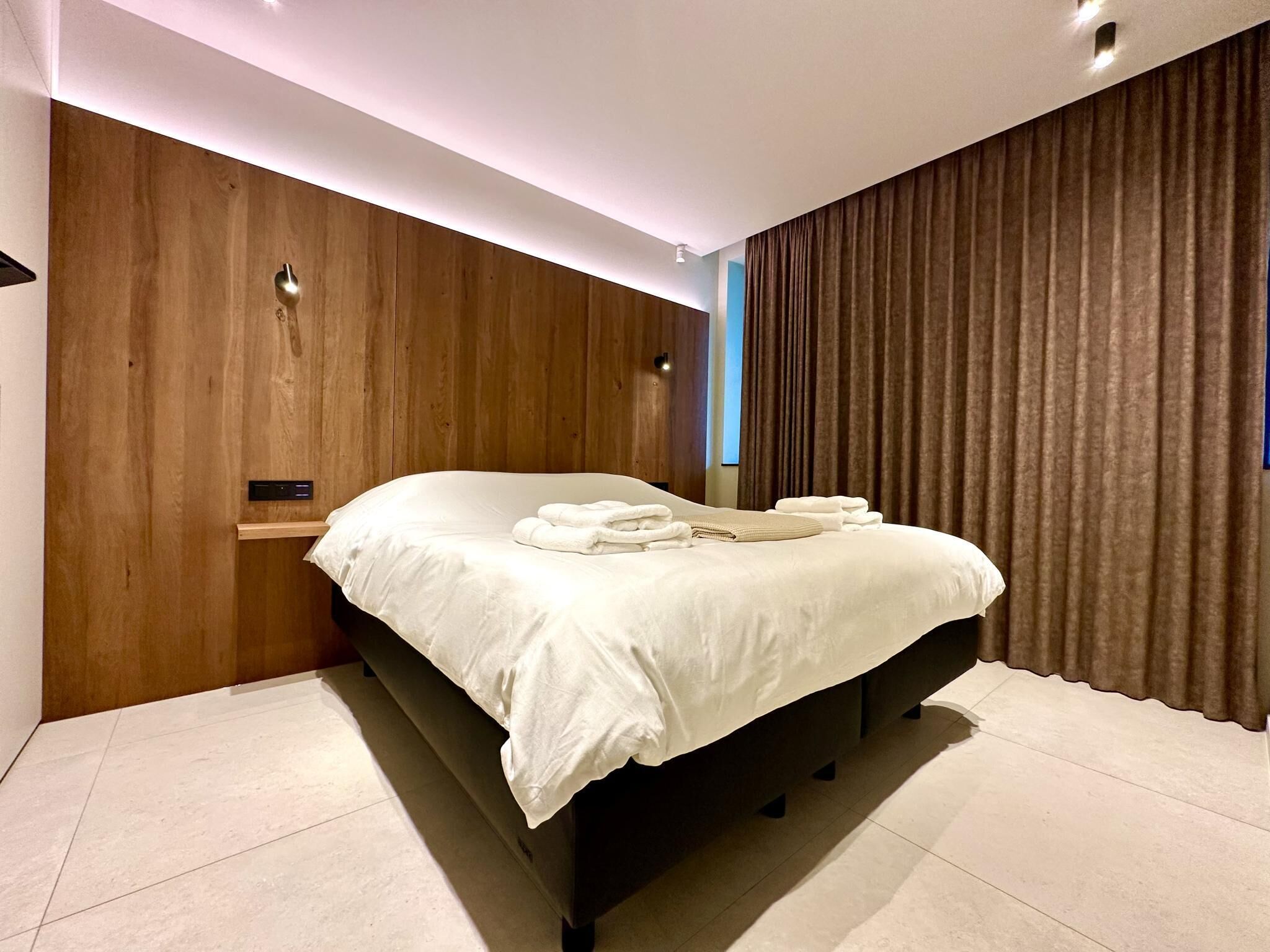 A modern bedroom with a large bed, wooden panel walls, and a curtain on the window.
