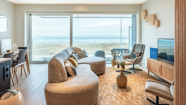 Ein modernes Wohnzimmer mit einer großen Couch, einem Couchtisch und einem Fernseher. Es gibt einen Balkon mit Blick auf den Strand.