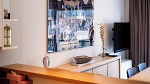 A modern living room with a large framed photograph on the wall, a flat-screen TV, a wooden coffee table, and a vase with dried plants.