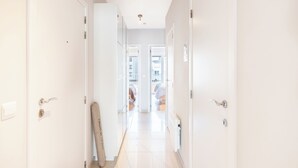 A bright, white hallway with a surfboard leaning against the wall, leading to a room with a bed and a window.