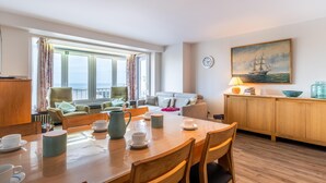 A well-lit room with a dining table set for four, a sofa, and a painting of a ship on the wall. - Cosy family flat with sea view (Koksijde)