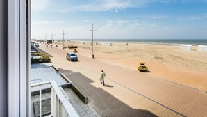 A beachside road with parked cars, a pedestrian, and beach huts. - Cosy family flat with sea view (Koksijde)