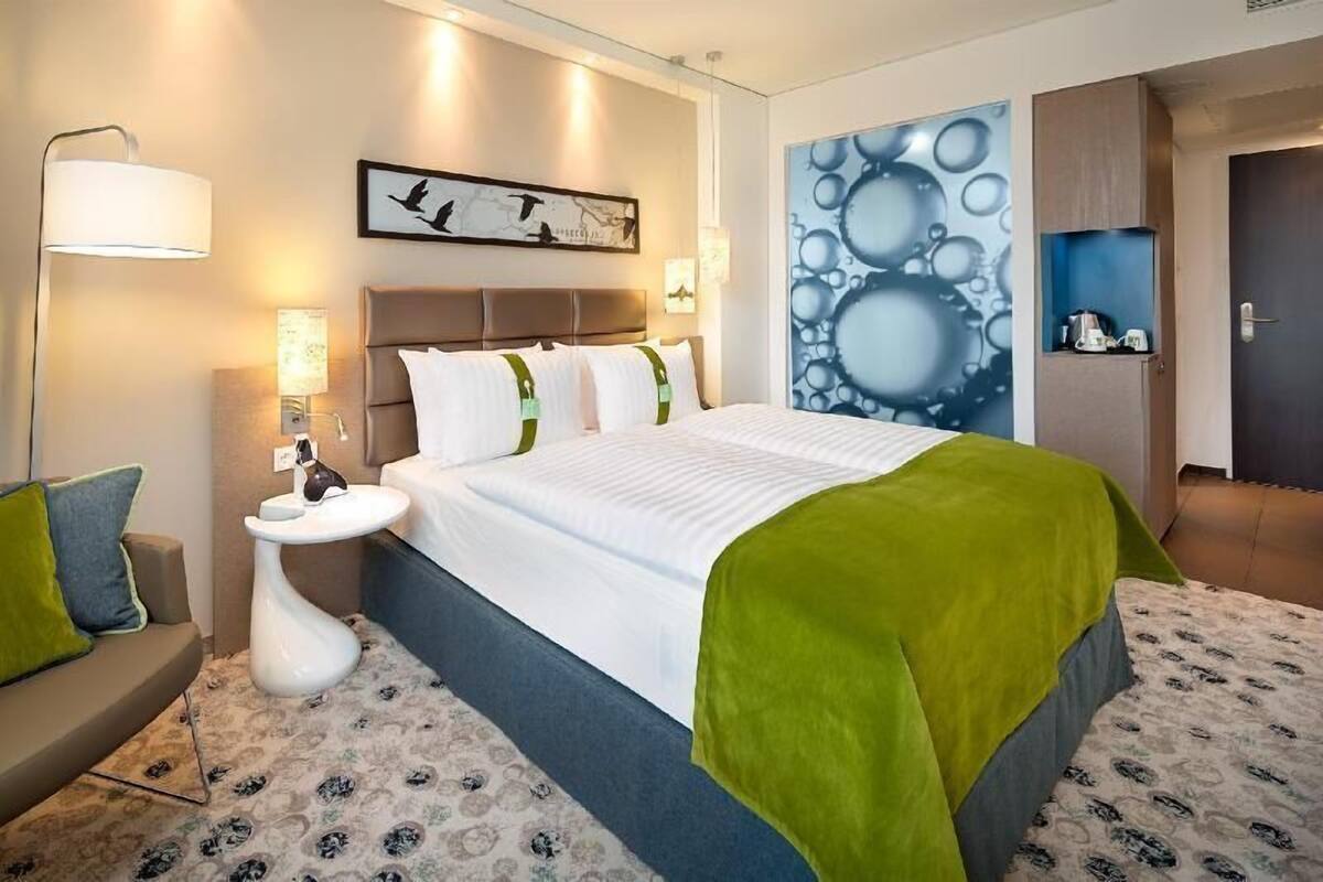 A hotel room with a bed, bedside table, lamp, and artwork on the wall.