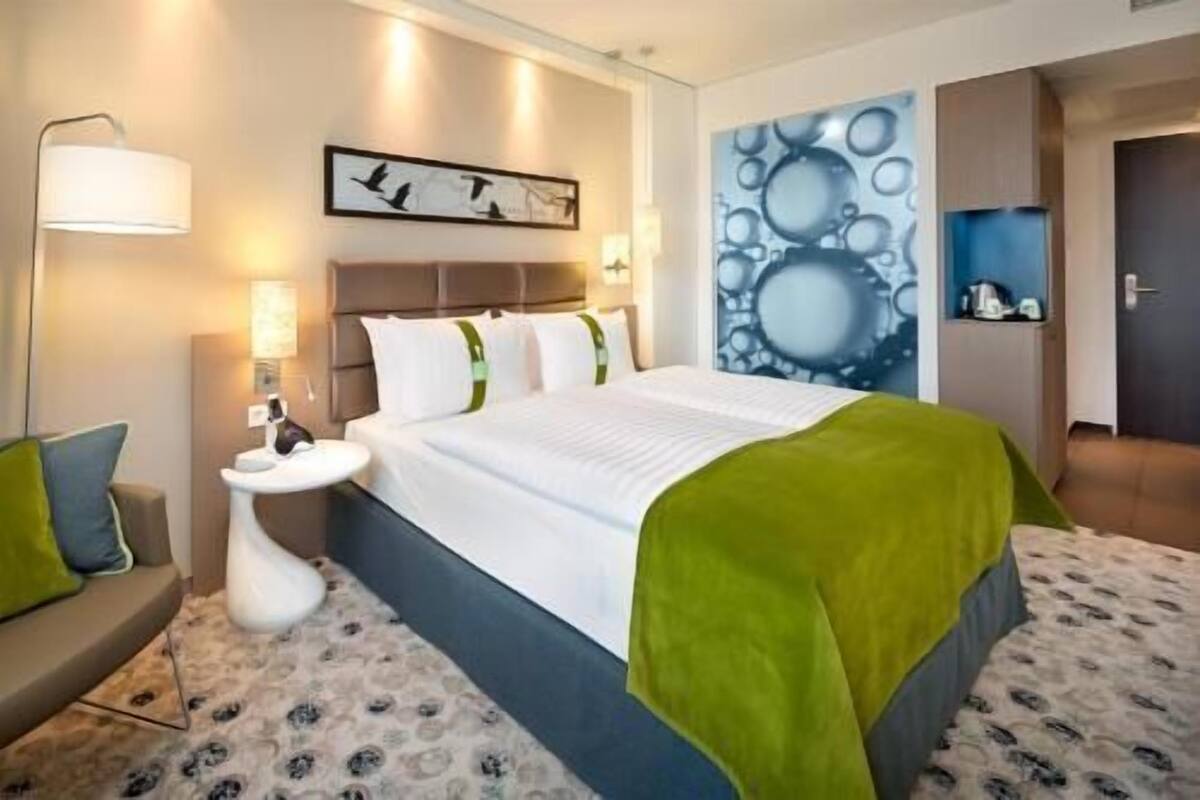 A hotel room with a bed, bedside table, lamp, and artwork on the wall.