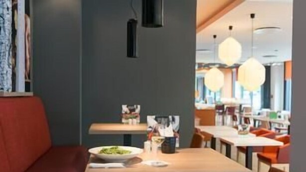 A modern restaurant interior with wooden tables, red seating, pendant lighting, and a view of the dining area. - Holiday Inn Berlin - City East Side (Berlin)