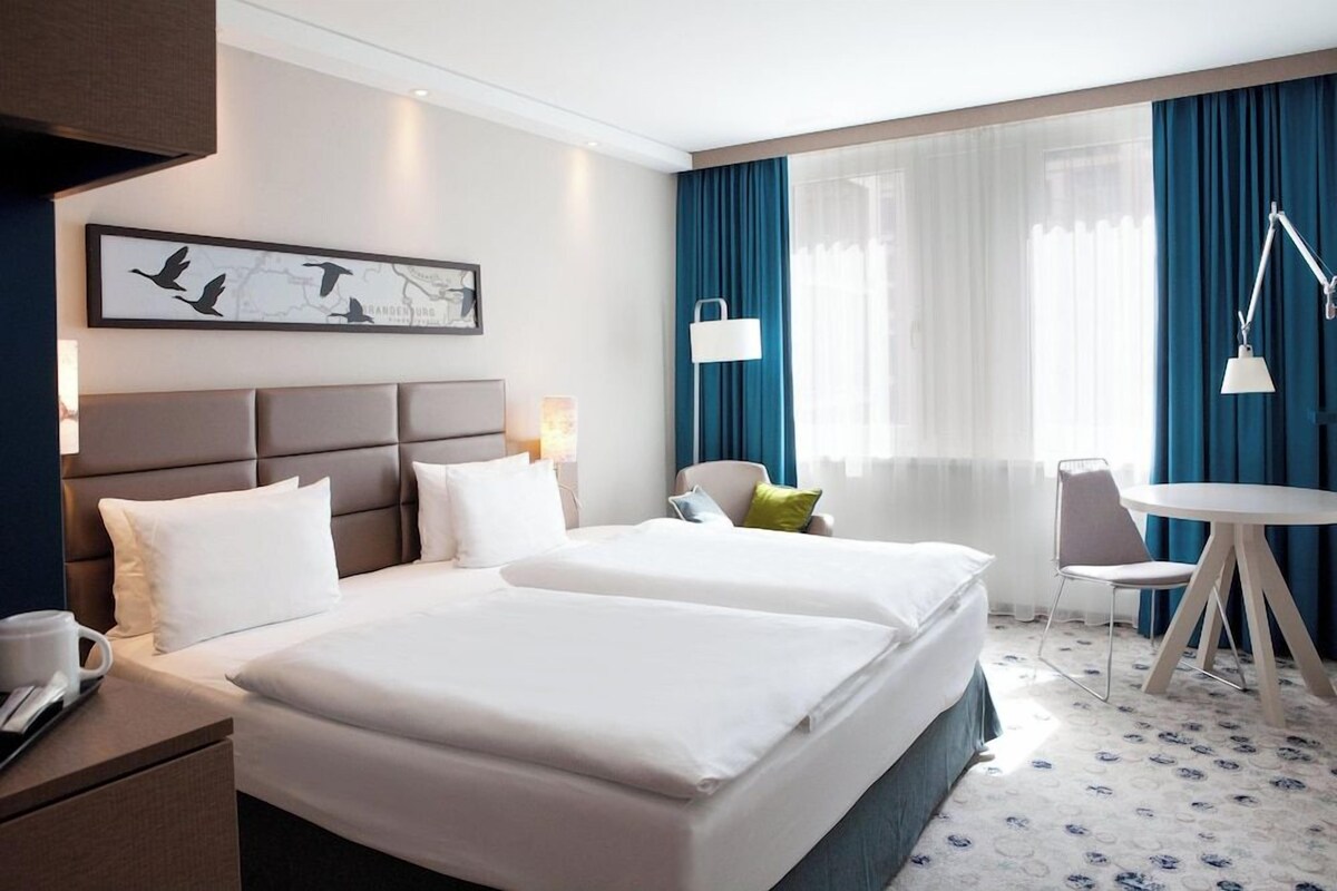 A hotel room with two beds, a desk, a chair, a table, and a large window with curtains.