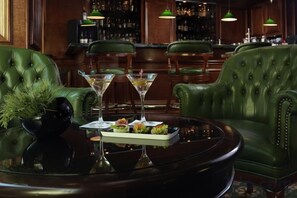 A bar area with green leather chairs, a round table with a tray of appetizers and two martini glasses, and a bar stocked with bottles. - NJV Athens Plaza (Athens)