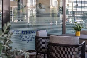 A seating area with wicker chairs and a table, a potted plant, and a view of a building with 'ATHENS PAA Lounge' written on the window. - NJV Athens Plaza (Athens)