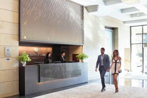 A reception desk with a modern design, a logo, and two potted plants. - NJV Athens Plaza (Athens)