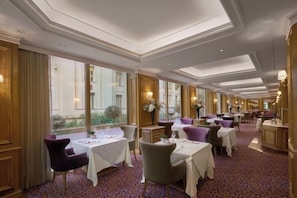 A hotel dining area with tables set for service, large windows, and a patterned carpet. - NJV Athens Plaza (Athens)