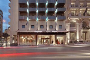 A multi-story hotel with a prominent entrance, large windows, and a covered area with a sign. - NJV Athens Plaza (Athens)