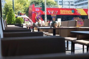 An outdoor seating area with tables and chairs, a view of a red bus, and a building with signage in the background. - NJV Athens Plaza (Athens)