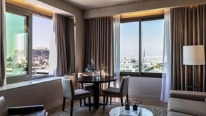 A hotel room with a dining table, two chairs, a sofa, a coffee table, and a view of a cityscape. - NJV Athens Plaza (Athens)