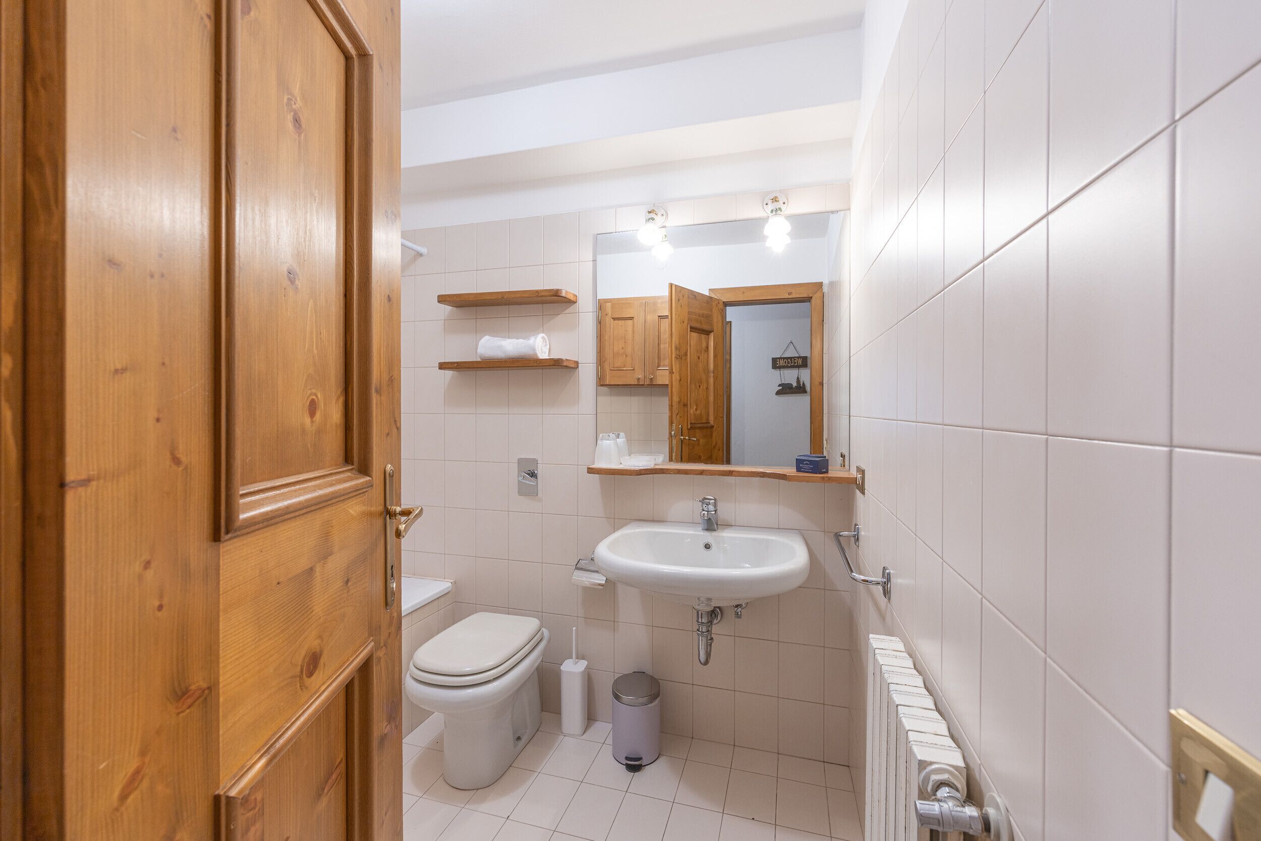 A bathroom with a wooden door, white tiles, a sink, a toilet, and a mirror.