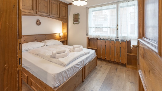 Quarto com estrutura de madeira, com cama, mesa de cabeceira, abajur e janela com cortinas de renda.