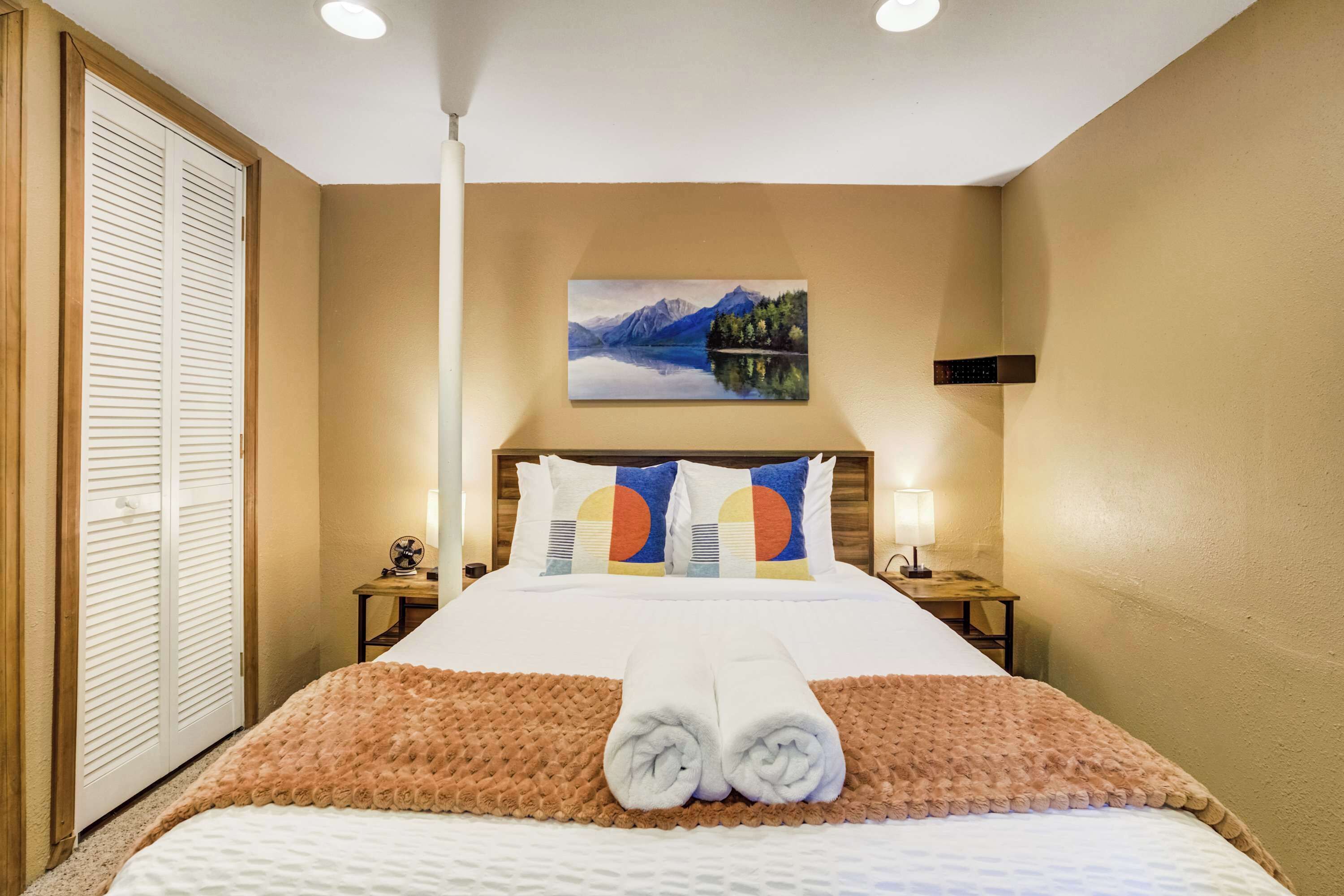 A hotel room with a bed, a painting of a mountain landscape, a wooden door, and a nightstand with a lamp.