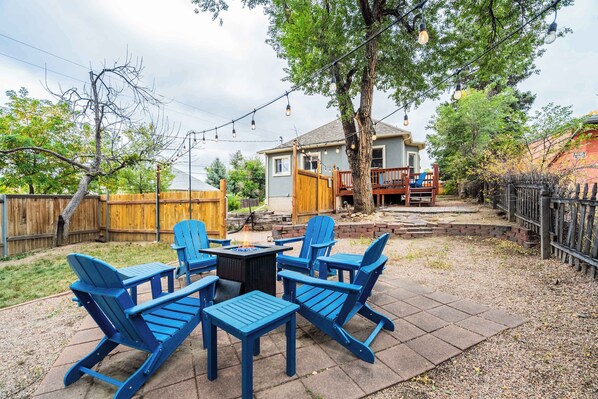 A backyard patio with four blue Adirondack chairs, a small table, and a fire pit.