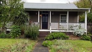 Ein zweistöckiges Haus mit Veranda, Eingangstür und einem kleinen Garten mit verschiedenen Pflanzen und Blumen.