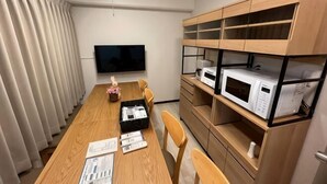 A room with a long wooden table, chairs, a flat-screen TV, and built-in shelves with appliances. - Core Royal-Vacation Rental (Kyoto)